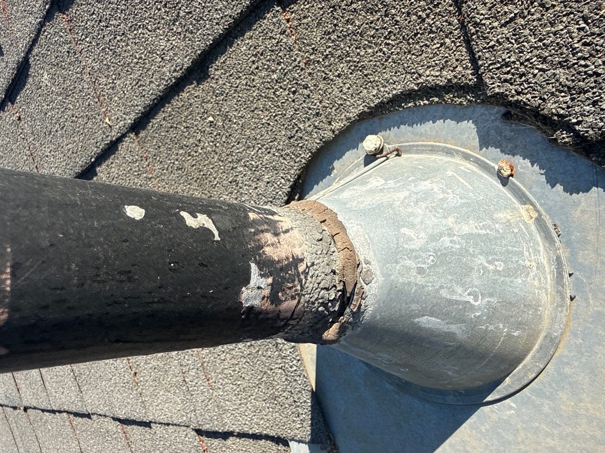 Cracked sealant at the metal collar where a black flue pipe passes through a shingle roof