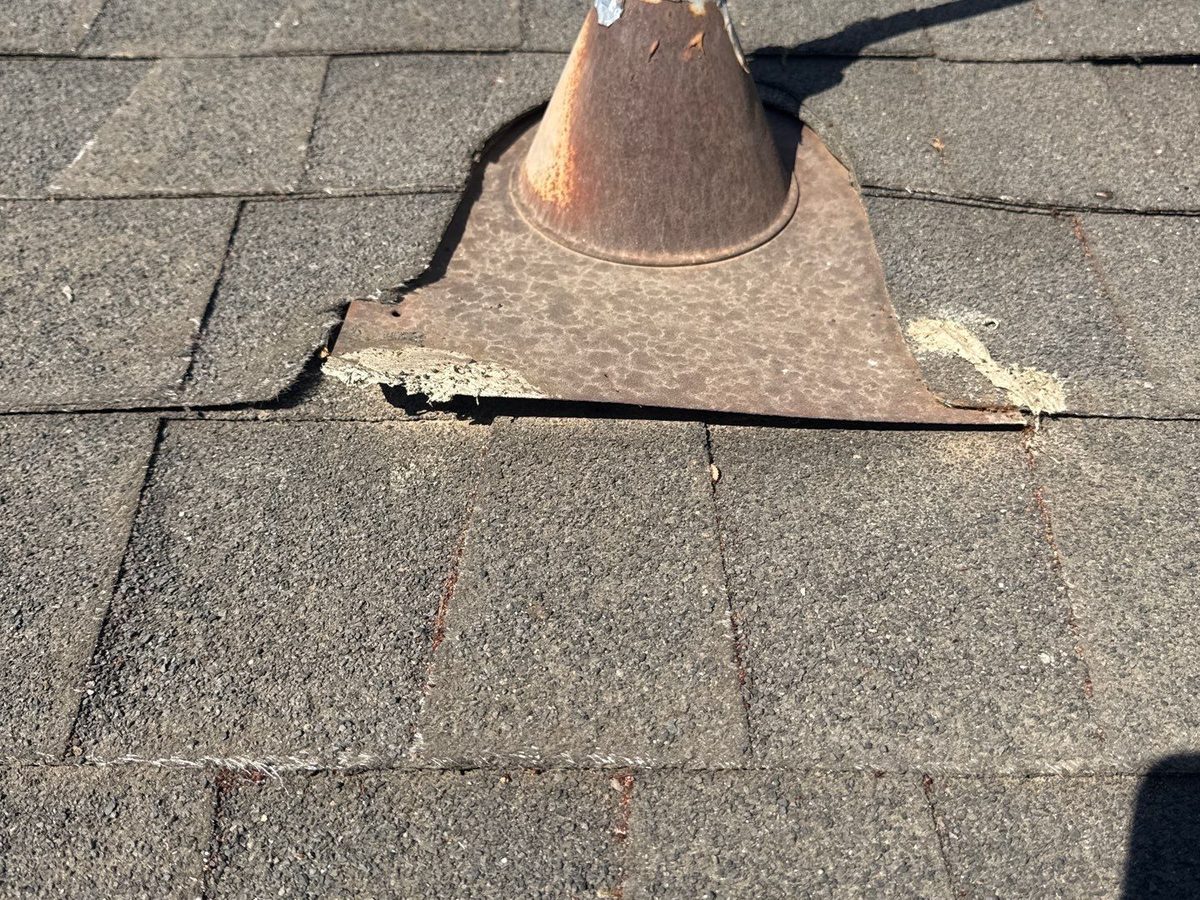 A rusted metal pipe flashing on a gray shingle roof, with the base lifting and the seal pulled away from the surrounding shingles